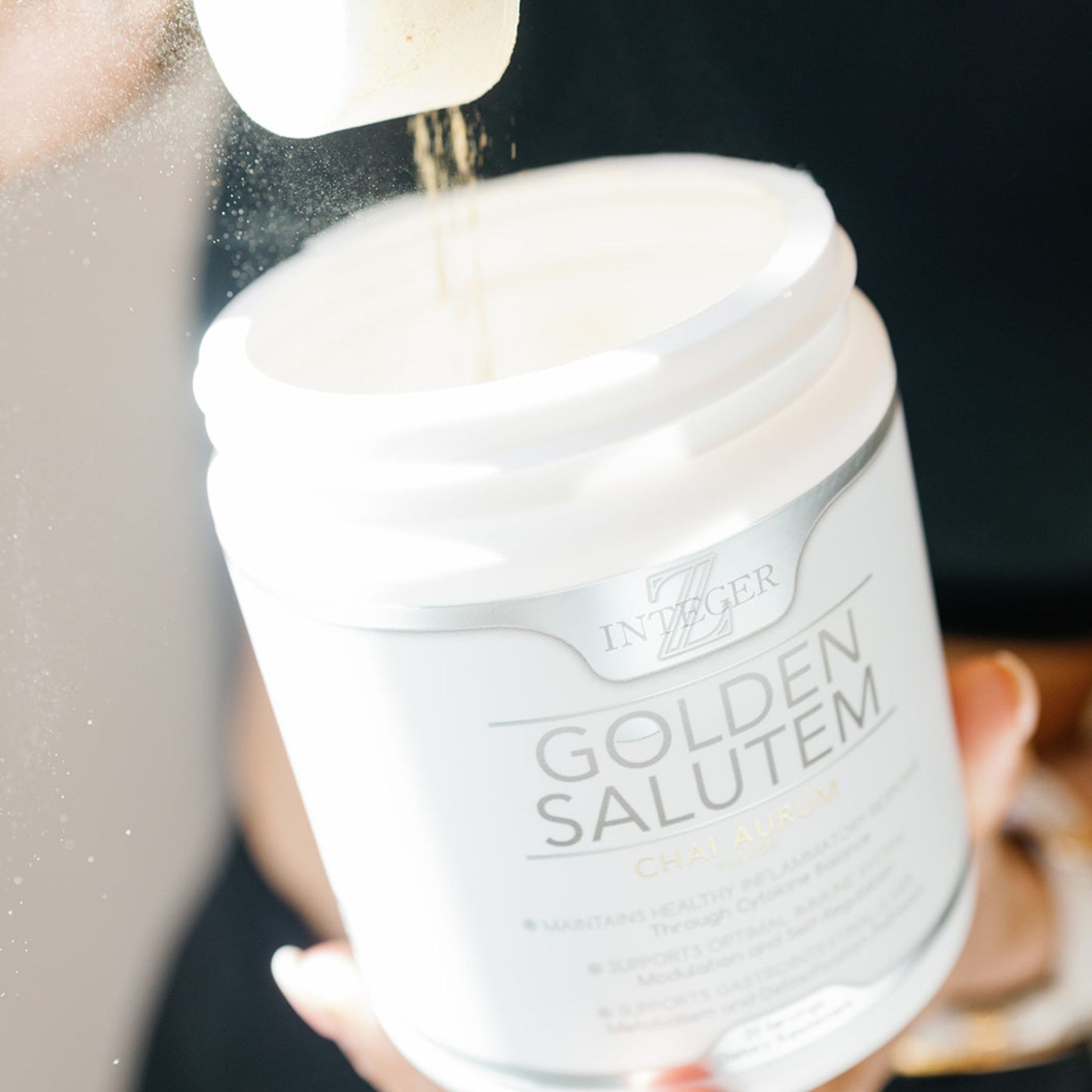 Open Golden salutem with scoop pouring powder back into tub being help by a woman with white nails and she is wearing black
