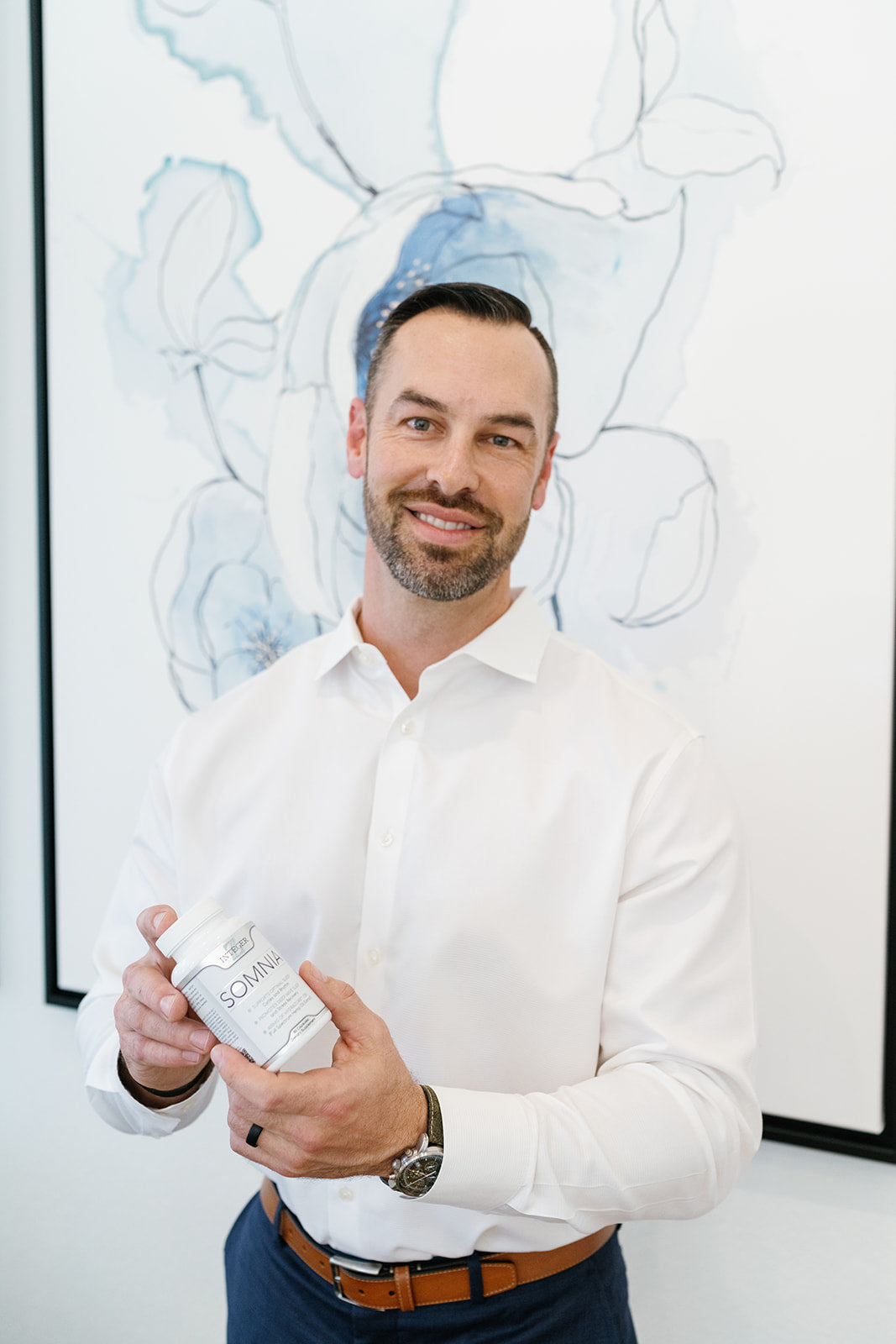 Owner a brunette tall man with a white shirt and black pants holding our sleeping product with blue and grey artwork behind him. 