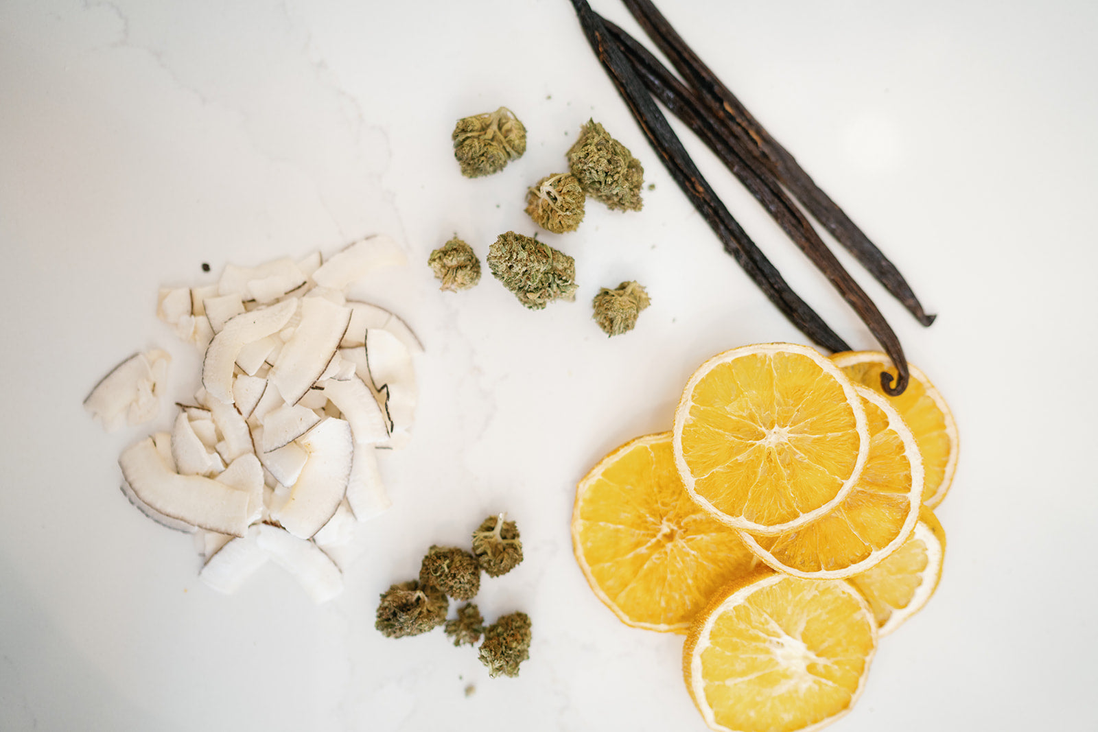Coconut chips, dried orange slices, vanilla bean pods and hemp flower laid out in a circle to show ingredients for a product