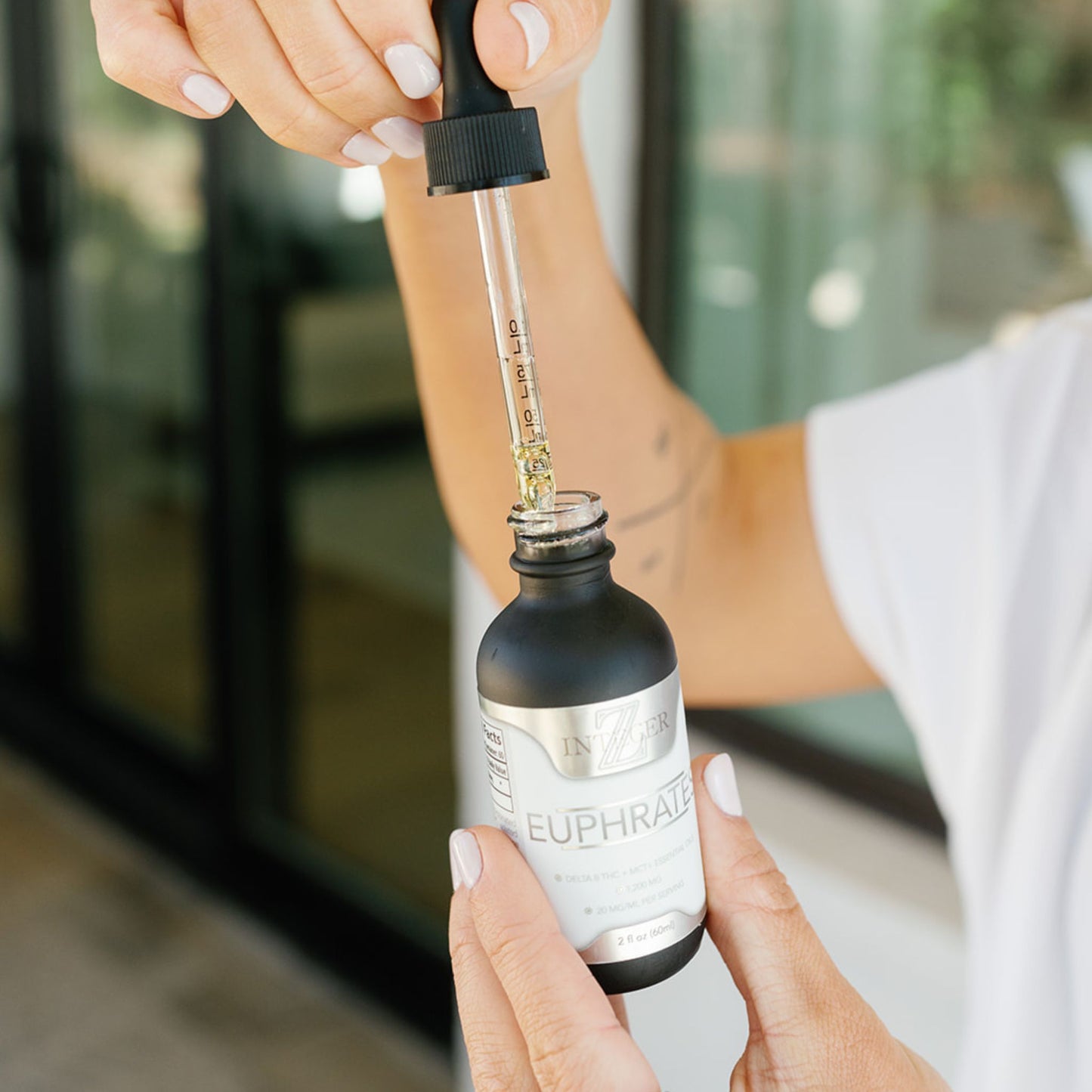 open euphrates bottle being held by a women with white nail polish  wearing a white shirt with windows to a home behind her