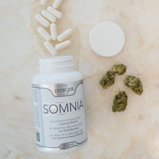 Somnia product with open cap and white capsules spilling out onto tan granite counter top with hemp flower laying next to bottle. 