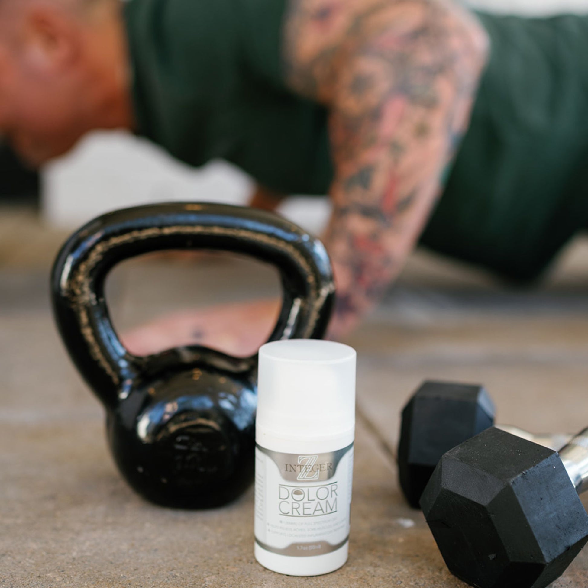 Dolor cream infront of a kettlebell and two dumbbells. Behind the weight there is a man in a green shirt with tattoos doing a push-up
