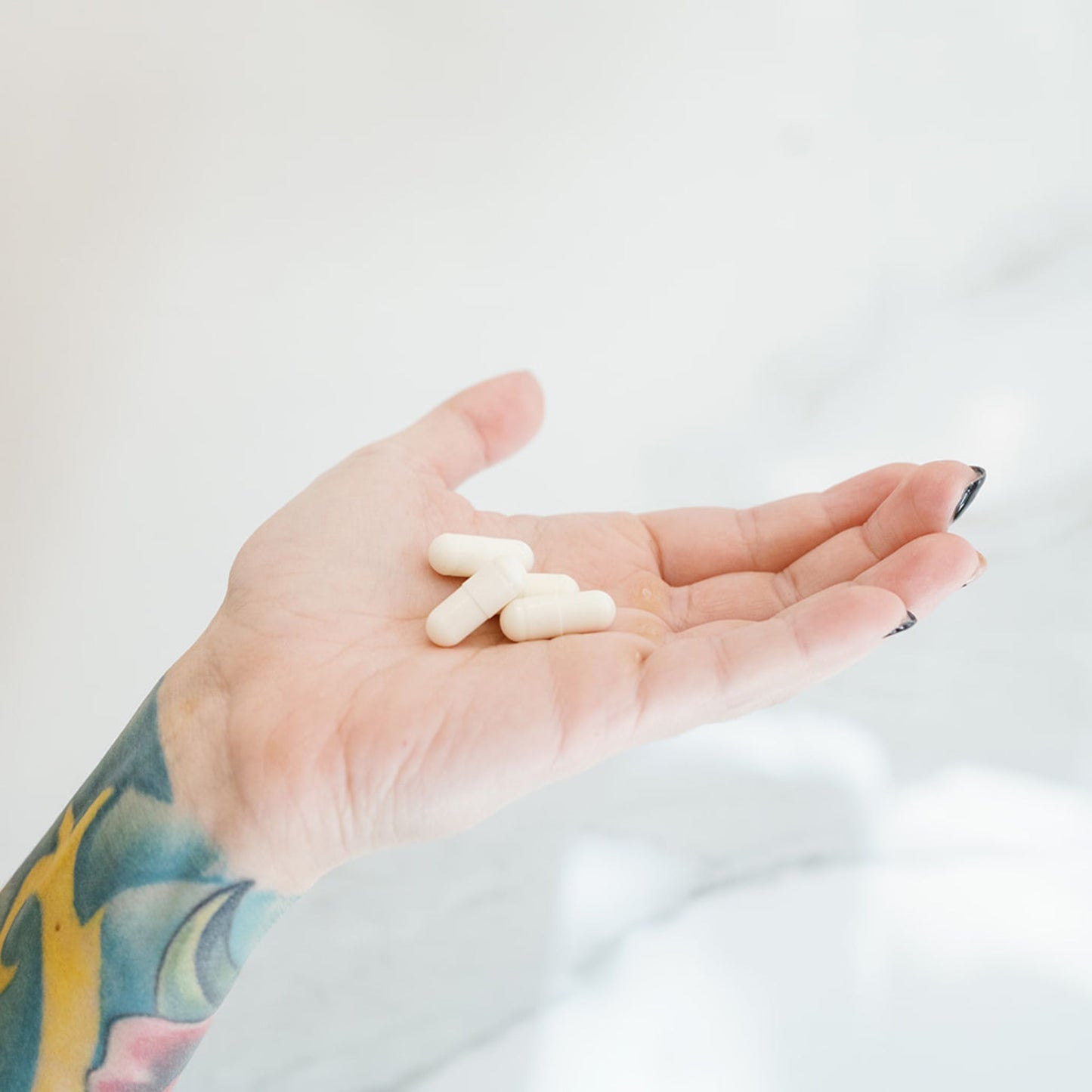 woman's hand with black nail polish and tattoos on forearm holding white capsules in palm.