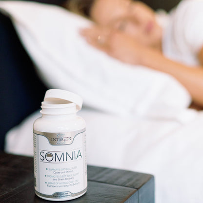 Somnia bottle with open cap on a black nightstand with woman behidn sleeping on white sheets and white pillow with white shirt on