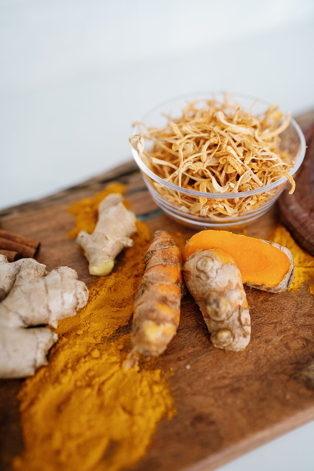 Wooden cutting board with Ginger root, turmeric cut open to show the vibrant orange color and mushroom root in a clear bowl
